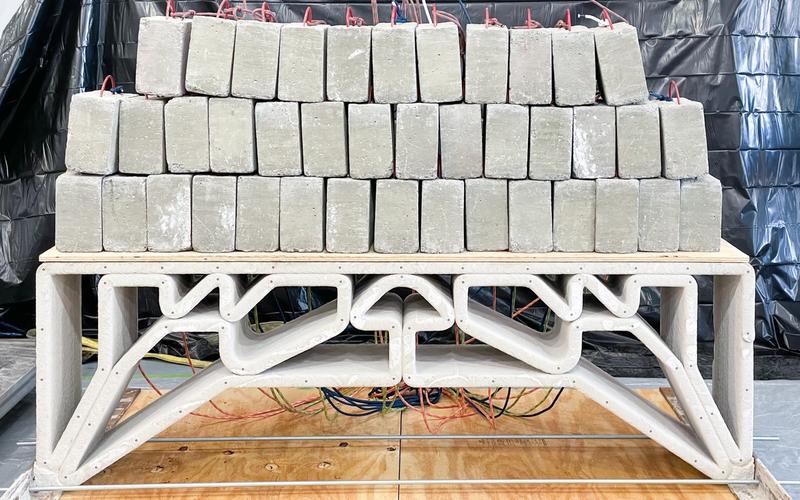 Stack of rectangular concrete blocks placed on top of a light-colored, 3D-printed concrete bridge-like structure with geometric supports, set on a wooden platform with colorful wires visible underneath.